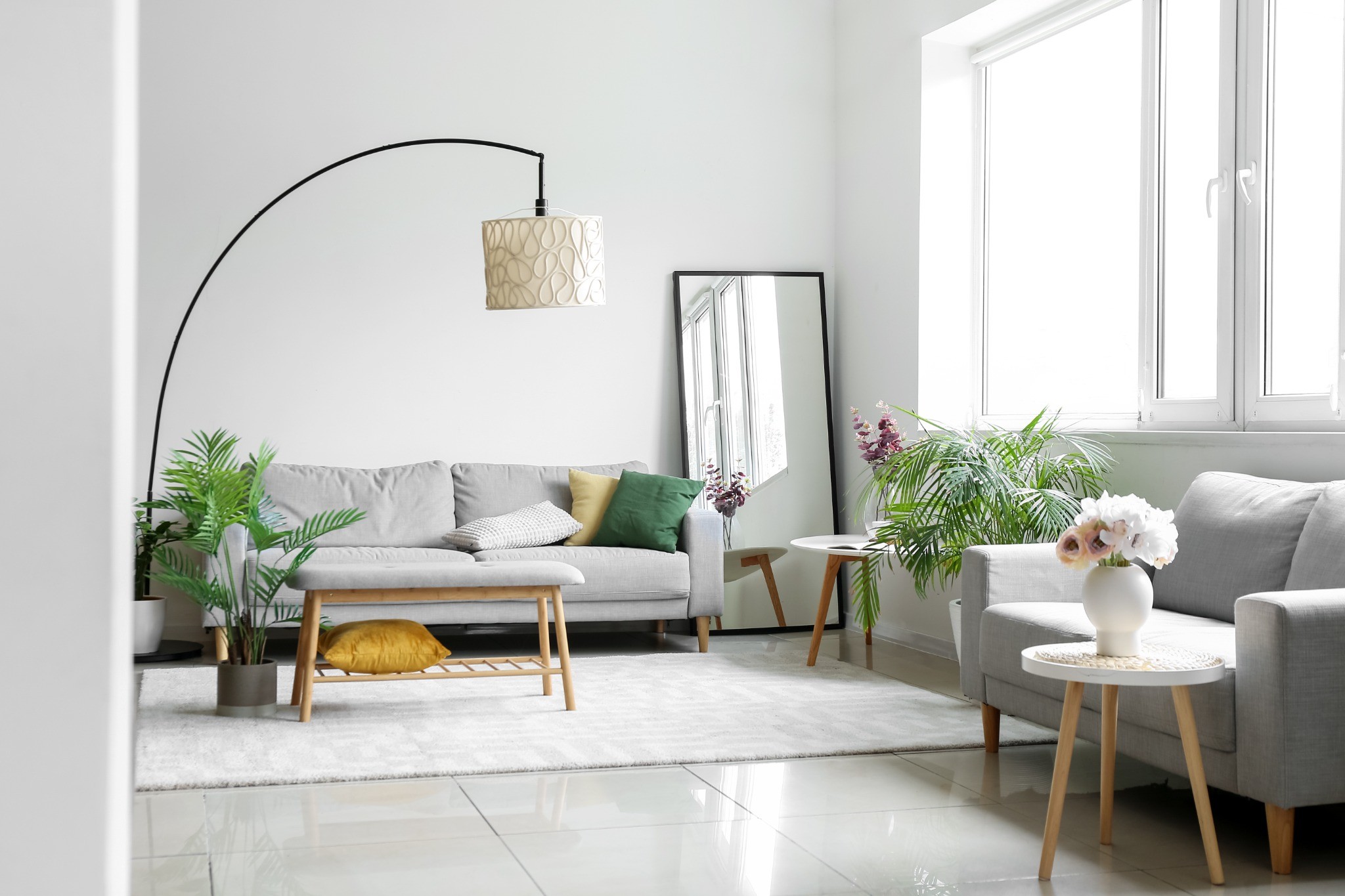 Salon moderne et lumineux avec un grand miroir rectangulaire posé contre un mur blanc, un canapé gris, une lampe à arc et des plantes vertes.