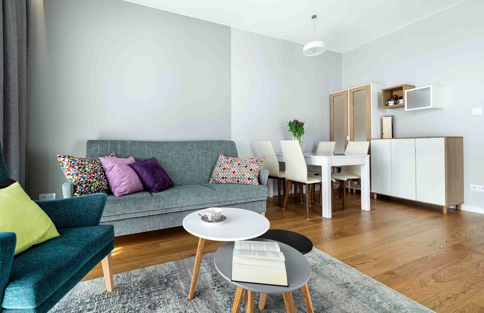 Vue d'ensemble d'un petit salon moderne avec un canapé gris, des coussins colorés et un coin repas blanc en arrière-plan.